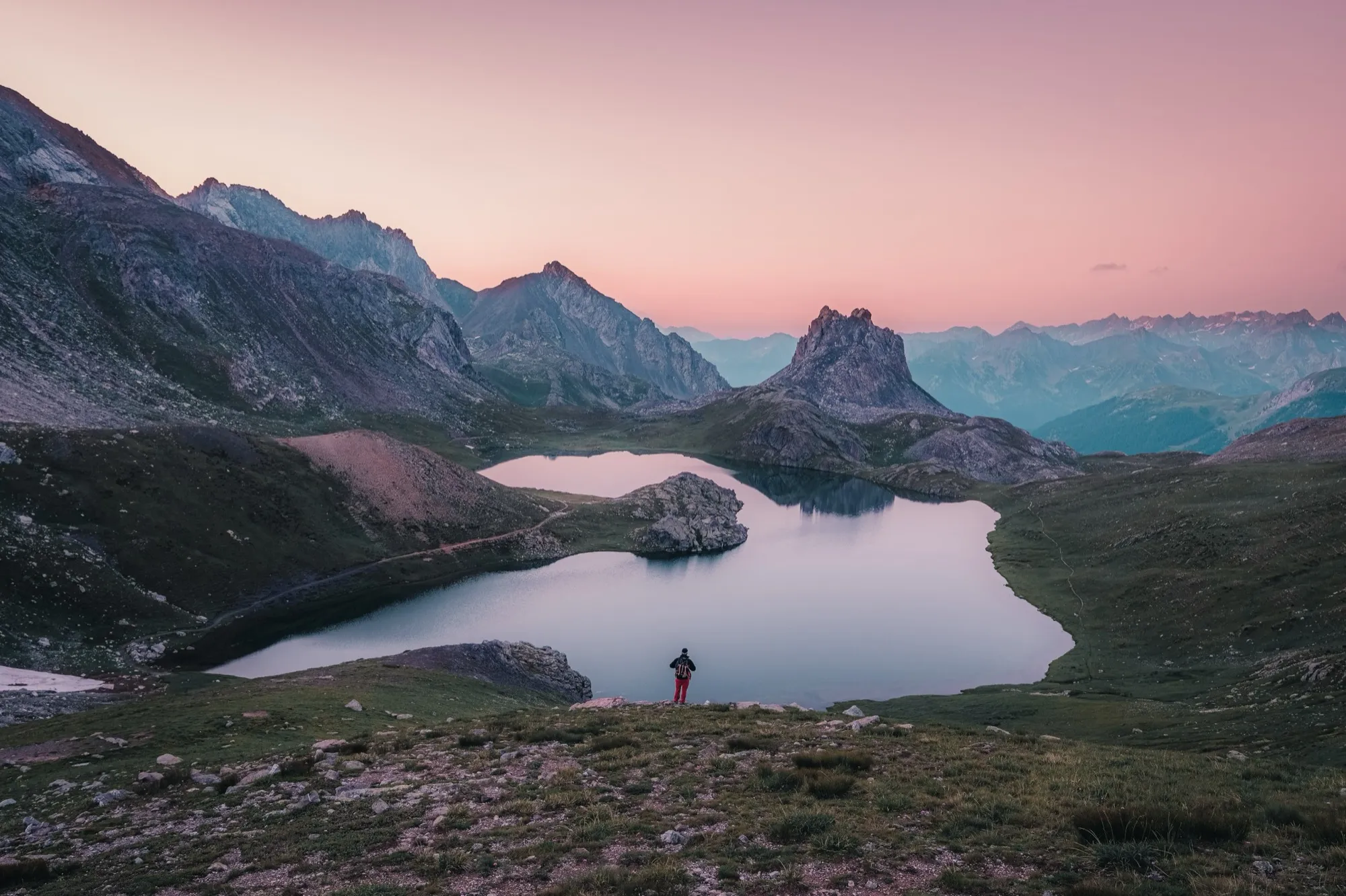Un randonneur face à un lac de montagne sous un ciel rose